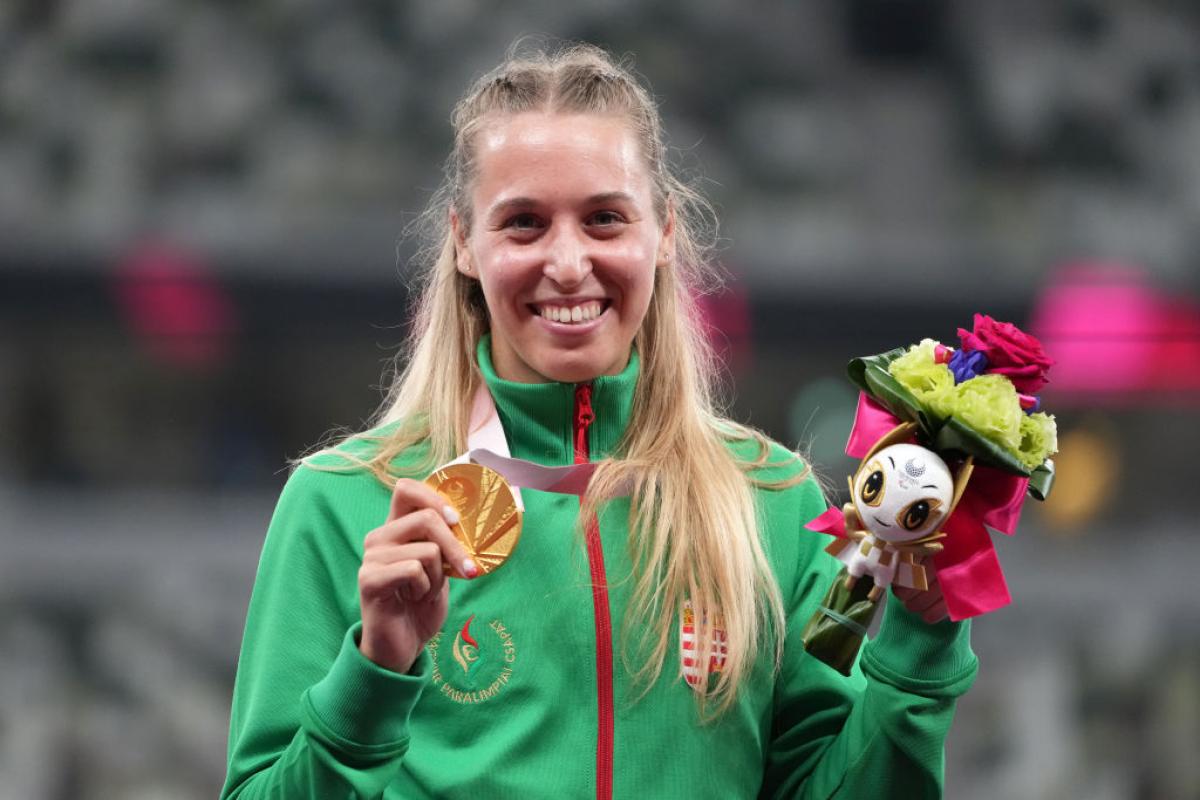 A female athlete smiles for a photograph while holding a gold medal with her right hand and a bouquet with her left hand.