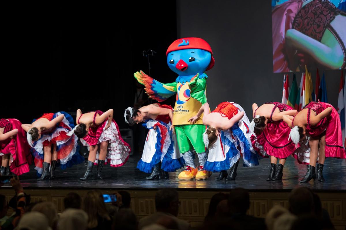 The mascot of the Virtus Global Games, which is a blue bird wearing a red cap, on a stage with seven women who are bowing.