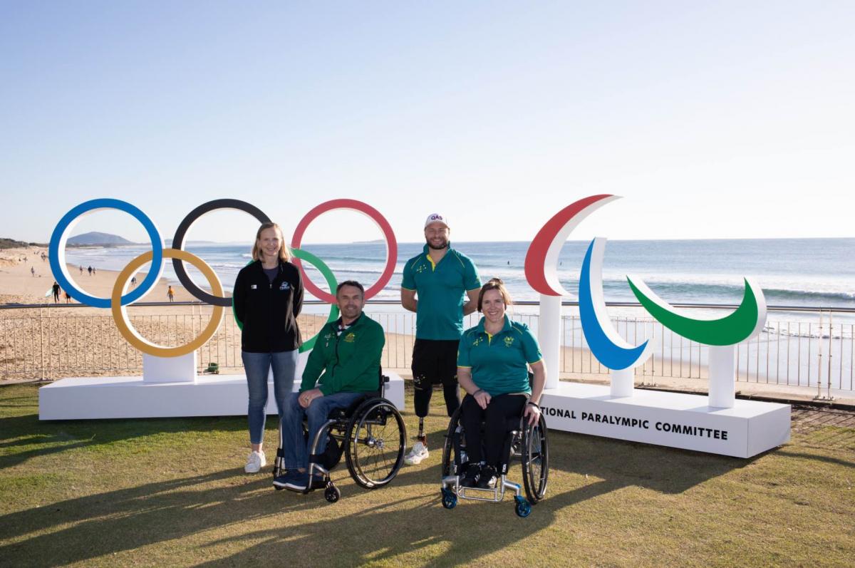 two wheelchair athletes and two athletes with prosthetics standing in front of the Paralympic and Olympic symbols 