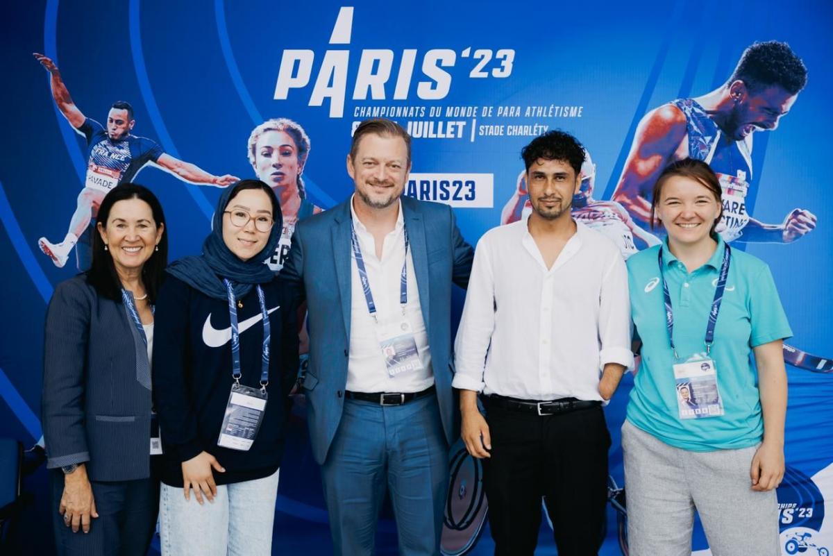 Five people pose for a group photograph in front of a Paris 23 Para Athletics World Championships banner