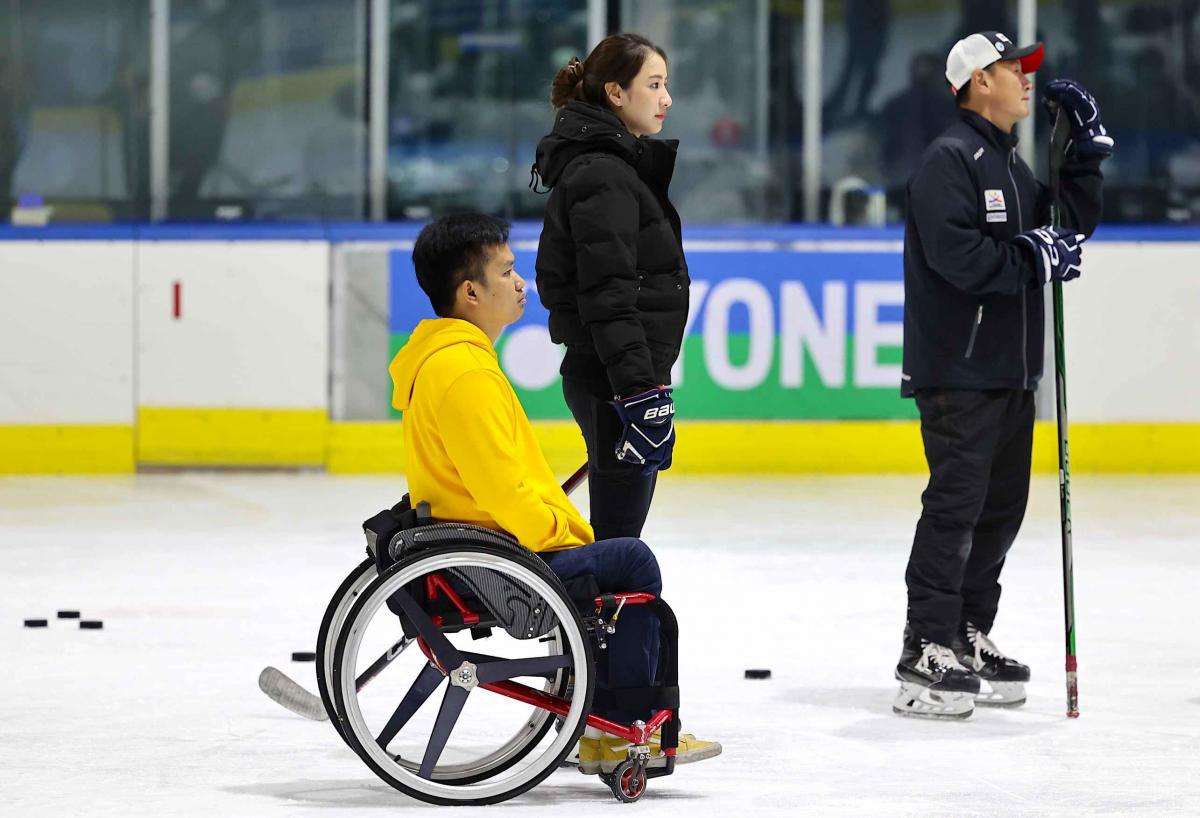 A man in a wheelchair next to a standing woman and a man on an ice rink