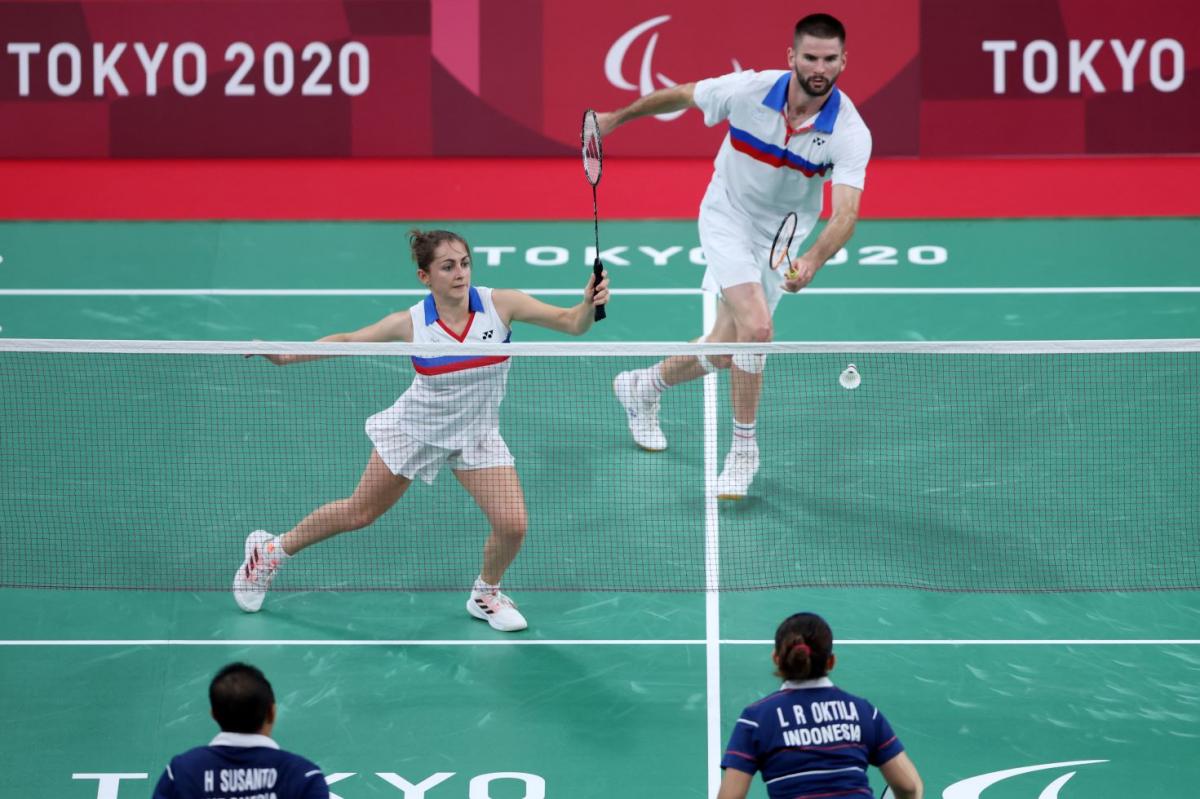 Photo shows a mixed doubles badminton match. A female athlete runs to a shuttle, in front of a male athlete.