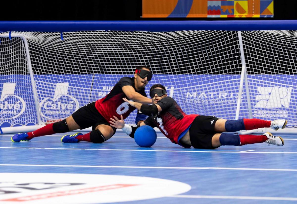 Two male athletes competing in goalball. They are blocking a blue ball in front of the net