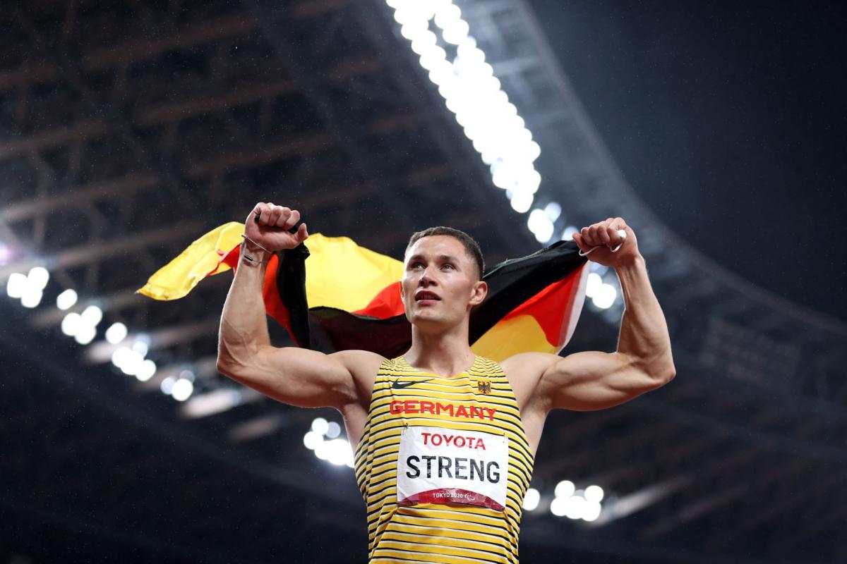 A male athlete holds the German flag with both hands at a stadium.