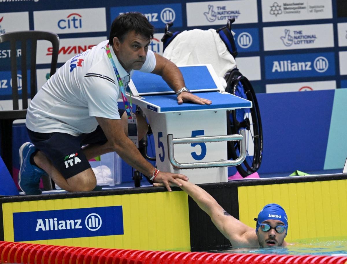 A male coach sits beside the pool and places his hand on a male swimmer's hand.