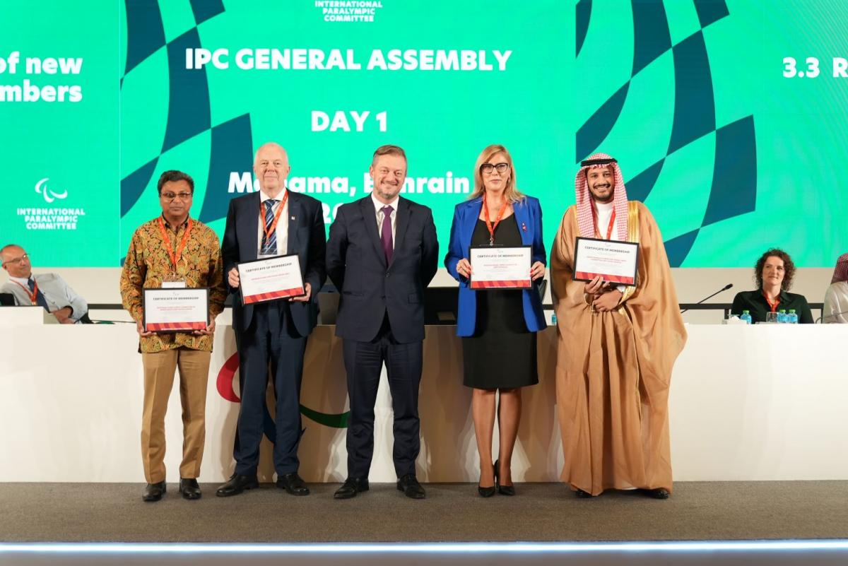Five people pose for a group photo in front of a green screen that says "IPC General Assembly".