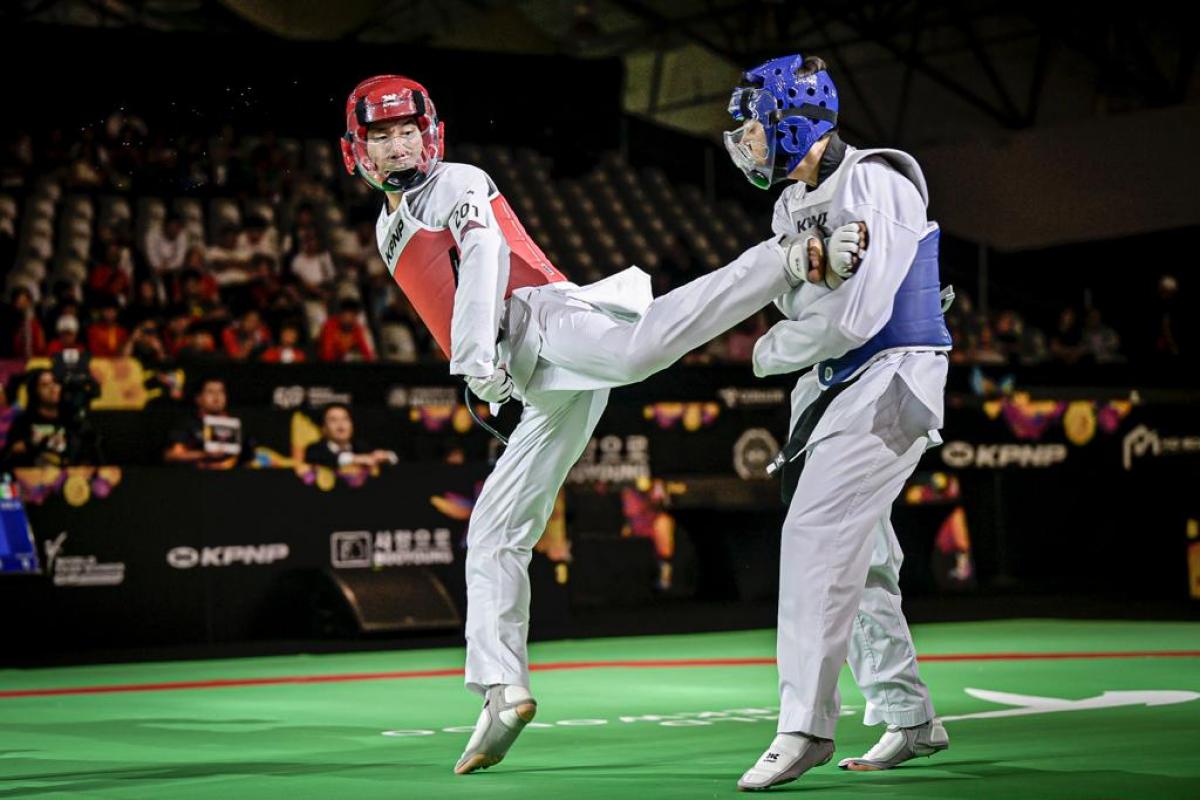 Two male Para taekwondo athletes fight in a match. An athlete wearing a red head gear kicks his opponent wearing a blue head gear.