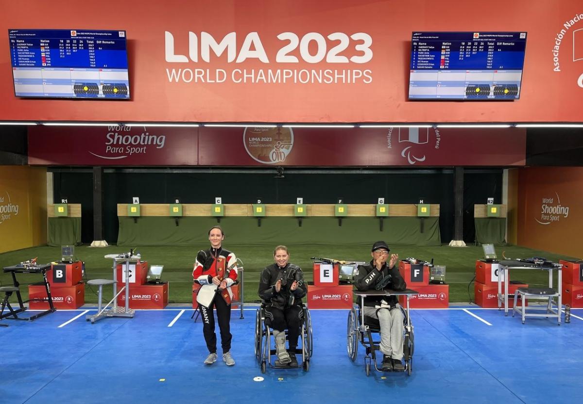 A standing woman next to a woman and a man in wheelchairs in a shooting range