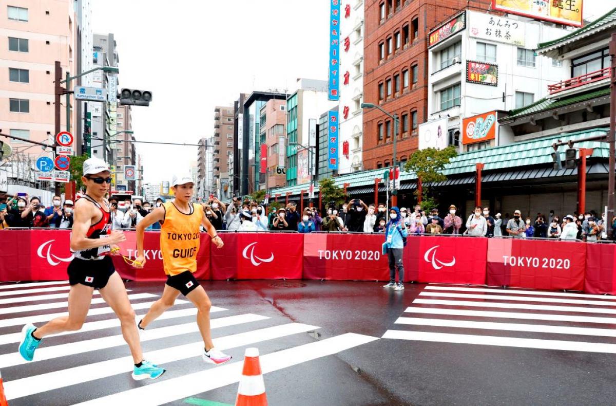 A male runner and his guide compete in the marathon at Tokyo 2020. People are taking photos as they compete.