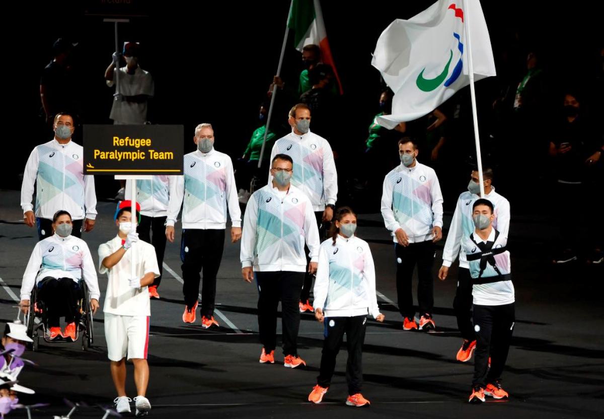 Ten athletes and officials take part in the athletes' parade at the opening ceremony of the Tokyo 2020 Paralympic Games. A volunteer is holding a sign that says "Refugee Paralympic Team" and a male athlete is carrying a flag of the Three Agitos.