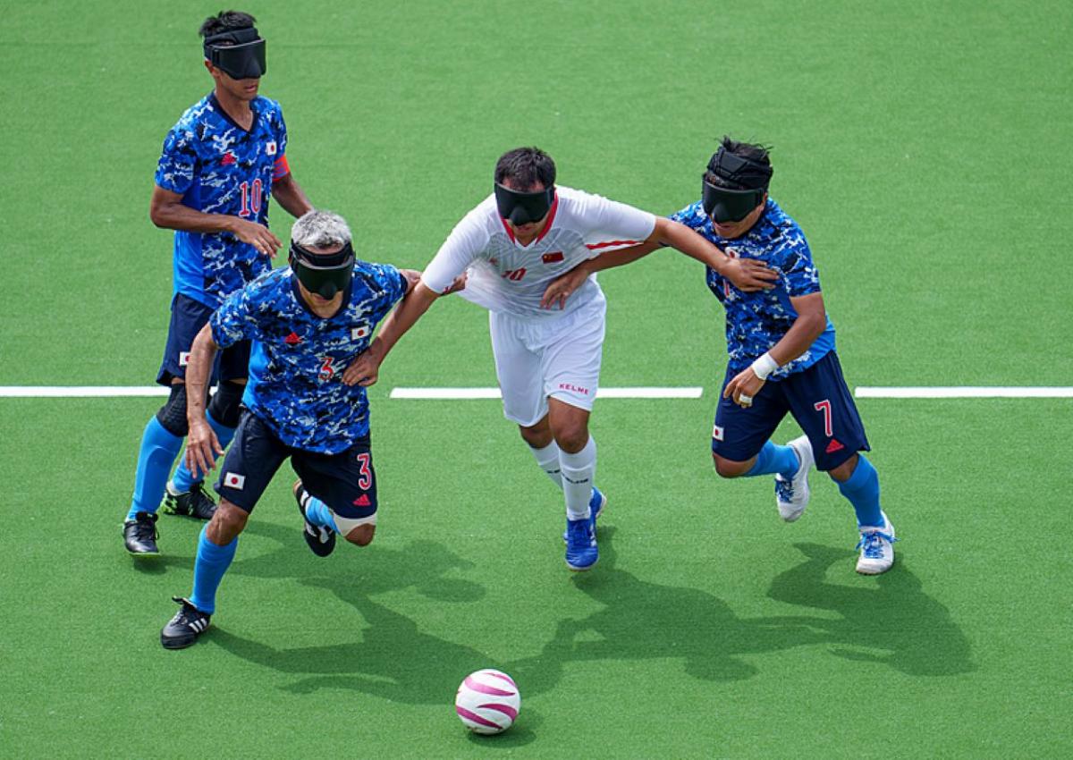 A blind football match between Japan and China at the Tokyo 2020 Paralympics. Three Japanese athletes wearing a blue jersey are trying to defend the ball from a Chinese athlete in a white jersey.