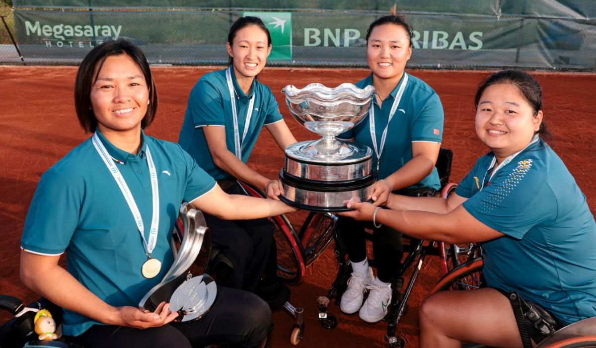 Four female wheelchair tennis athletes pose for a photograph. They are holding a silver trophy.