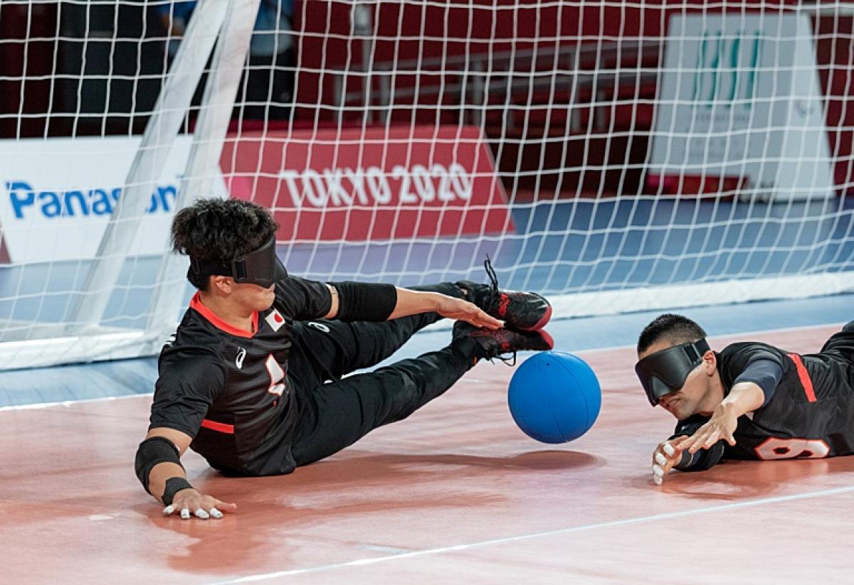 Two male goalball athletes are blocking the ball in front of the goal. They have their hands on the court.