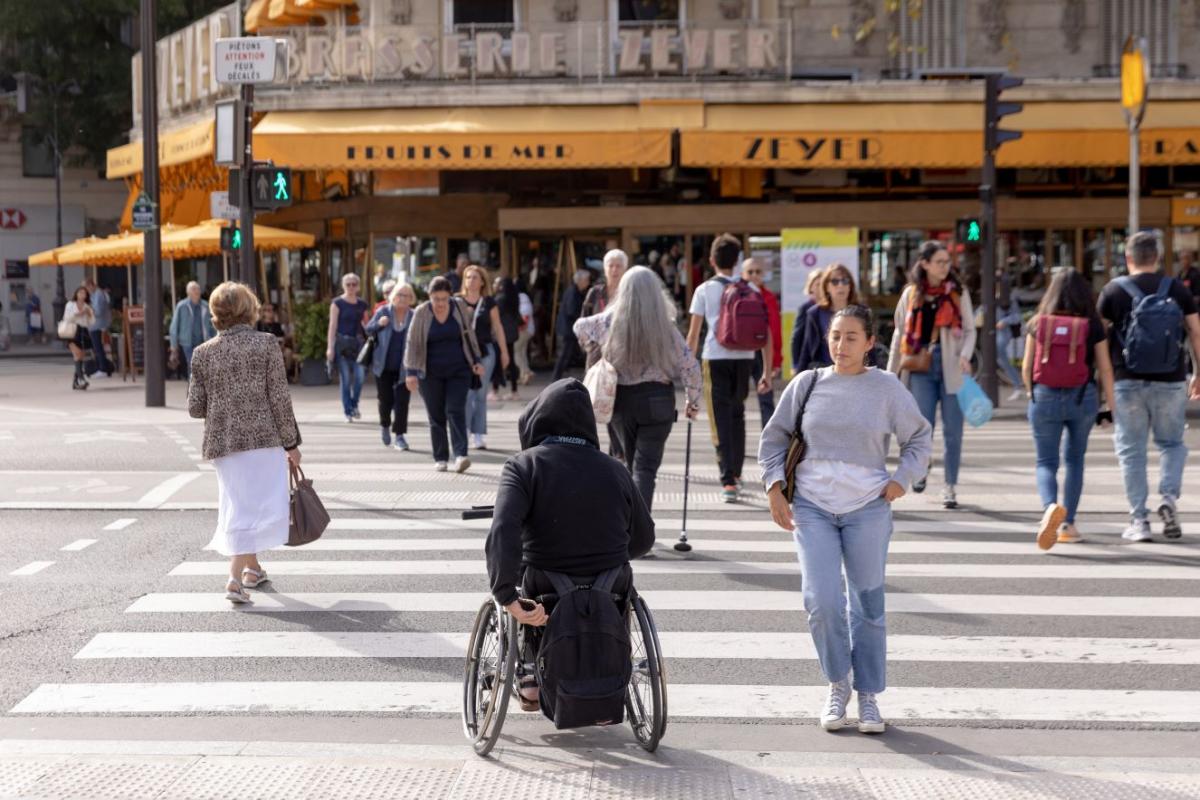 Many people cross a sidewalk in Paris. There are people in a wheelchair and people using canes.