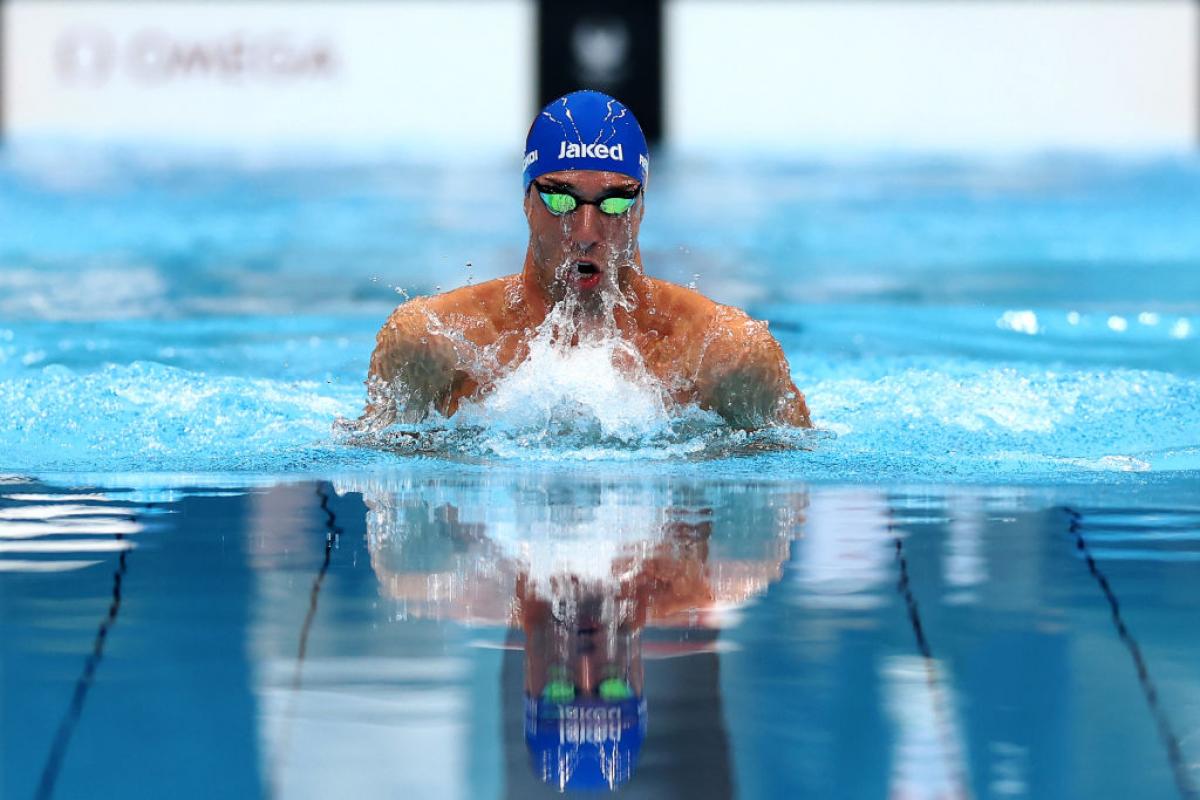 The head and shoulders of a swimmer in a blue cap and googles emerges from the water with a splash
