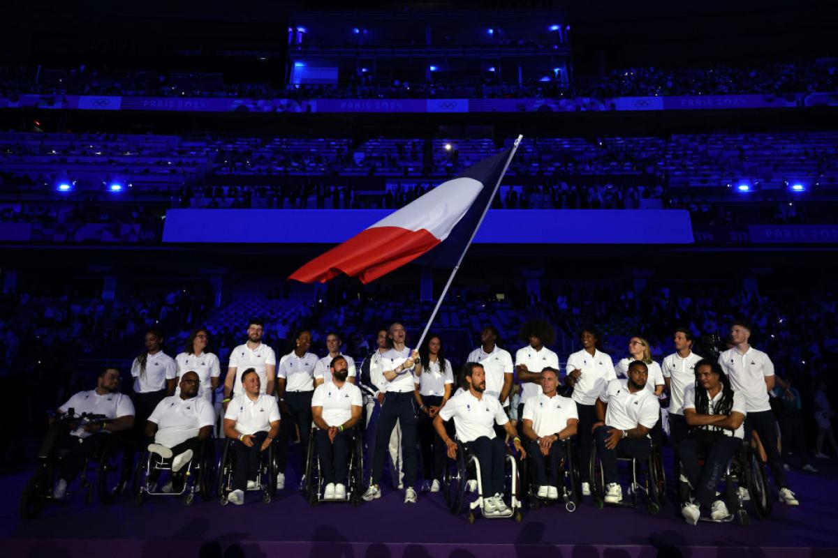 About 20 Paralympic athletes taking part in the Closing Ceremony of the Olympic Games. The female athlete in the centre is waving the French flag.