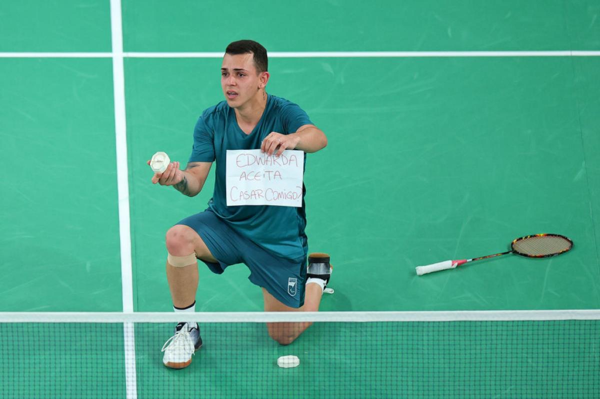 Man in blue tee and shorts kneels on the badminton court holding a ring