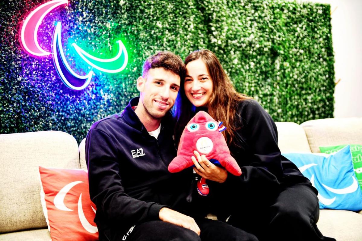 Man and woman face camera dressed in blue with Agitos in neon lights behind them holding the Paris 2024 mascot