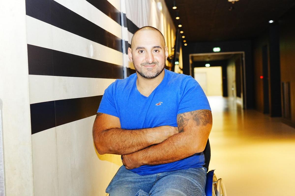 Man seated in blue t-shirt smiles to camera with arms folded