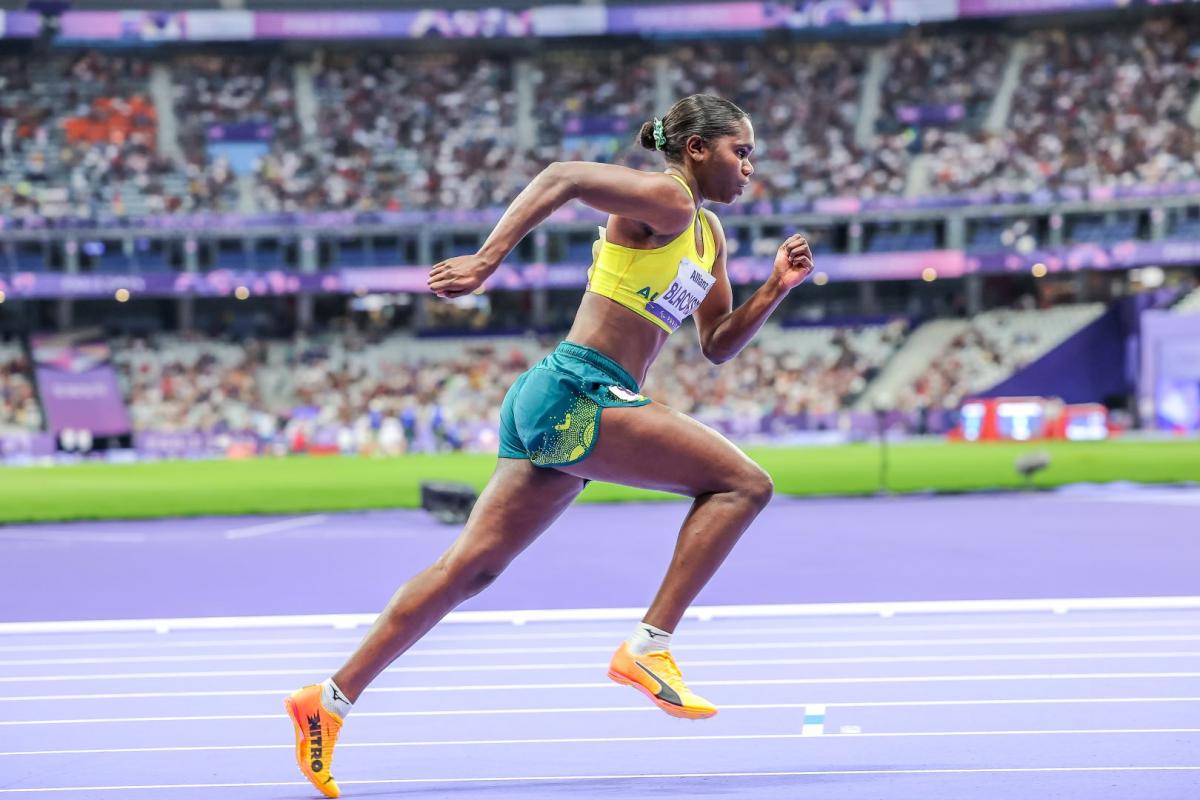 An Australian female athlete with a yellow vest and green shorts runs at the Stade de France athletics stadium