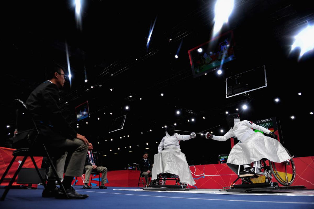 Two athletes in wheelchairs fence. They wear white uniforms and black face shields, their swords are crossed.