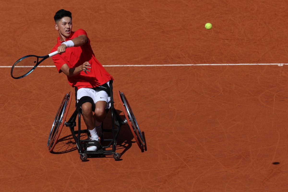 Against an orange earth background a man in a red tee and a wheelchair reaches a racket out as a yellow tennis ball flies