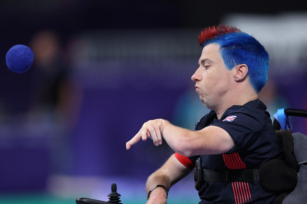 A man in a wheelchair with red and blue hair throws a blue boccia ball