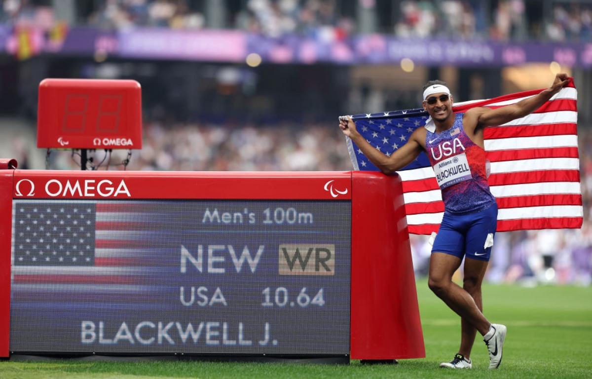 A man in a Team USA uniform holds up an American flag and stands next to an LED screen that shows his new world record time