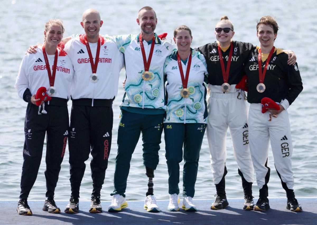 Six athletes stand on a podium next to water, two with silver medals, two with gold medals, and two with bronze medals