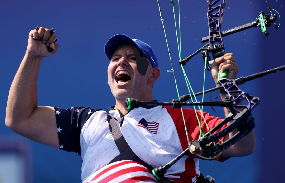 A man raises both arms in celebration, holding his archery bow in one hand