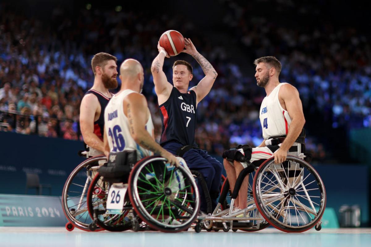 A man in a wheelchair holds a basketball above his head, surrounded by three other wheelchair basketball players