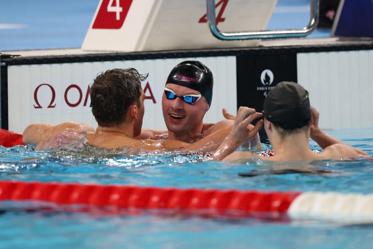 Three men are in a swimming pool, two are congratulating a third, who is wearing a swim cap and goggles.