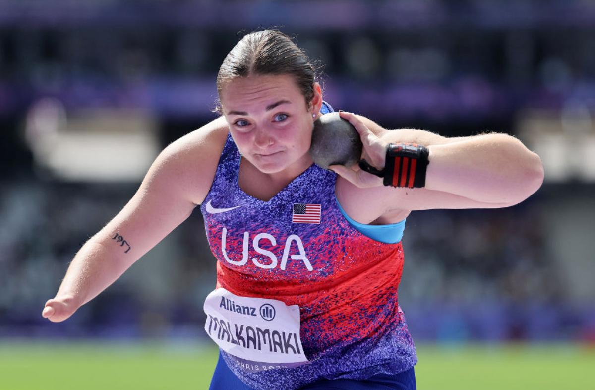 A woman in a Team USA uniform holds a shot put up by her neck to prepare to throw it