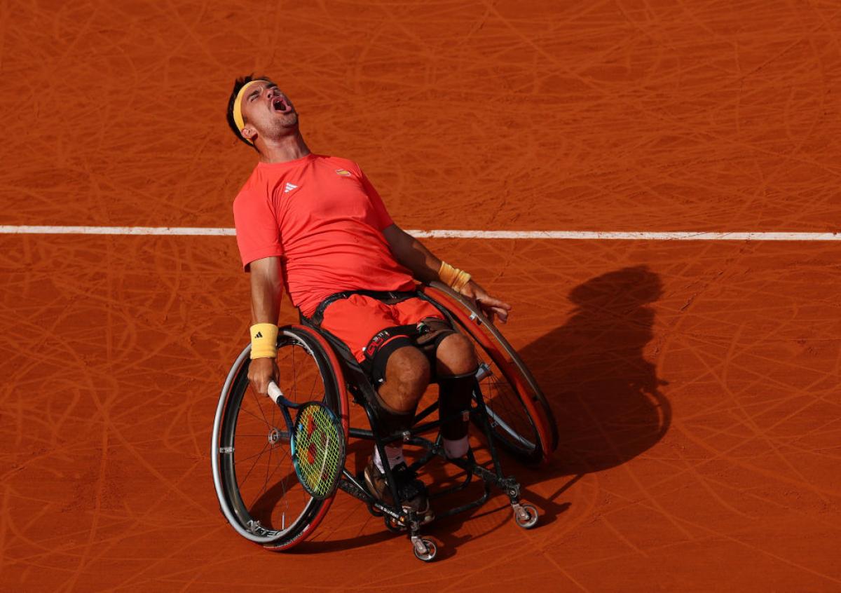 Man leans back in his wheelchair holding a tennis racket closing eyes and screaming, against a backdrop of a clay tennis court