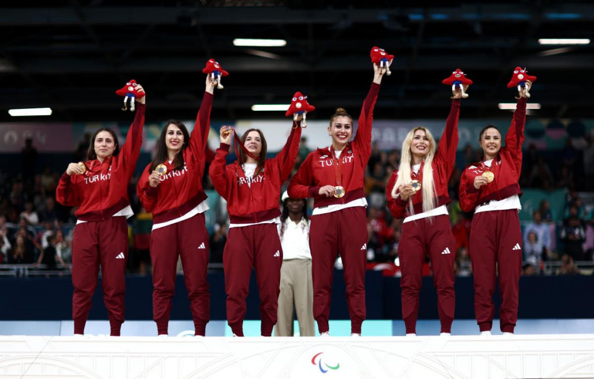 six women in red Turkiye uniforms hold up the Paris 2024 mascot and their gold medals with smiles on their faces