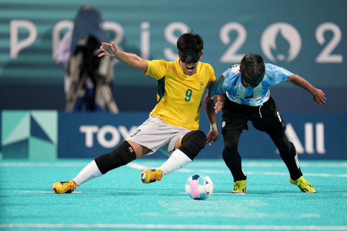 Two blind football players wearing black eyeshades, one in a yellow Brazil jersey and one in a blue and white Argentina jersey, compete for the ball