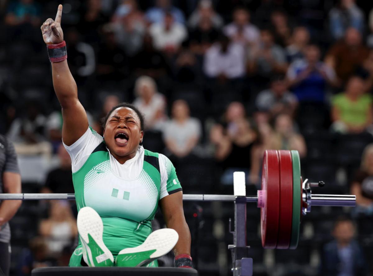 Woman in white and green sport outfit is seated with her hand in the air in a Number 1 sign, she scrunches her face to scream, with powerlifting weights behind her