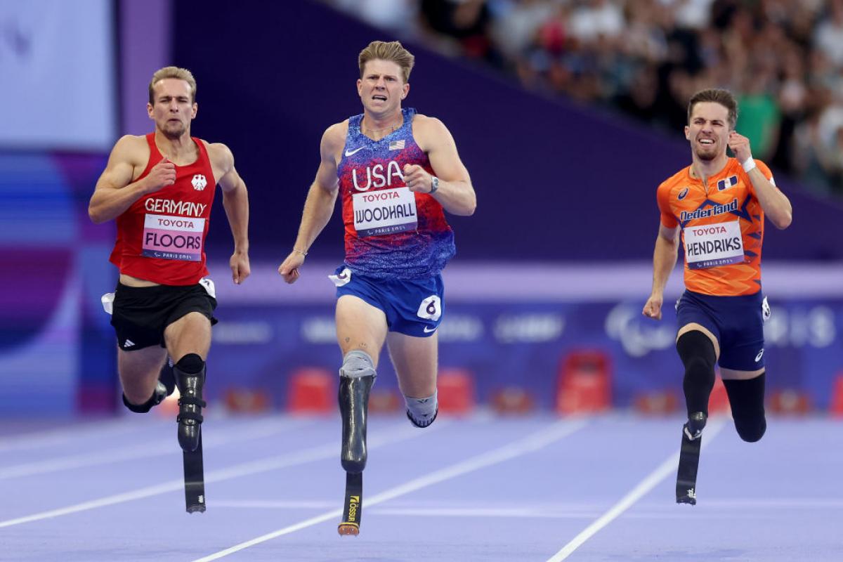 Three men with running blades race against each other, with the man in the middle, in a Team USA uniform, ahead of the other two
