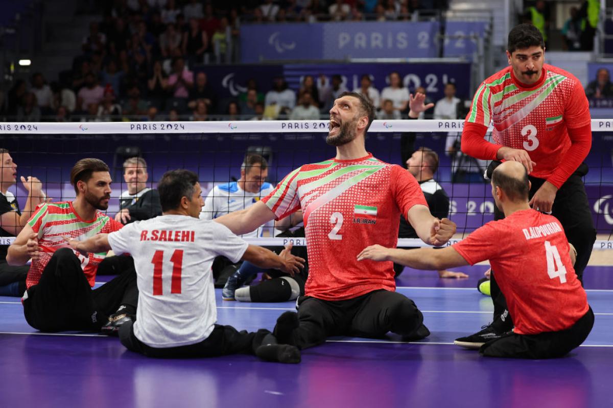 Five players wearing Team Iran uniforms yell in celebration during a sitting volleyball match