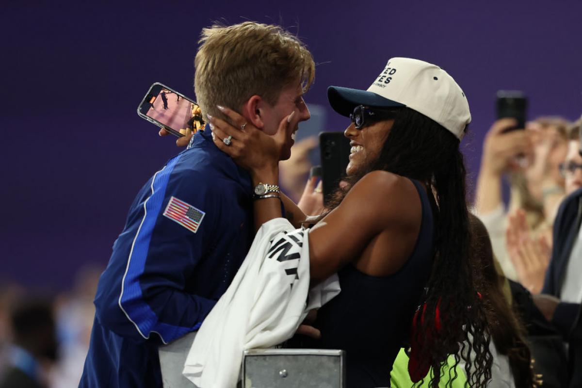 A woman in the crowd in a United States baseball hat puts her arms around a man in a Team USA jacket, both smiling
