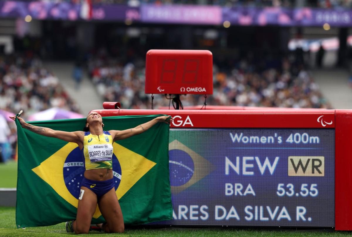 A female Brazilian athlete celebrates with her country's flag in front of her new world record of 53.55 on the screen