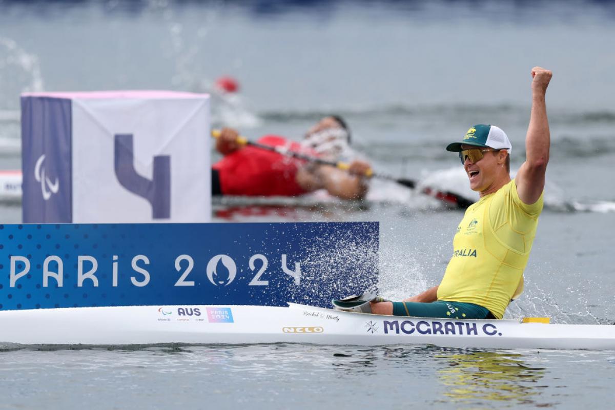 An Australian male rower wearing a yellow shirt, sunglasses and a green hat, celebrates a rowing win by punching the air in his boat