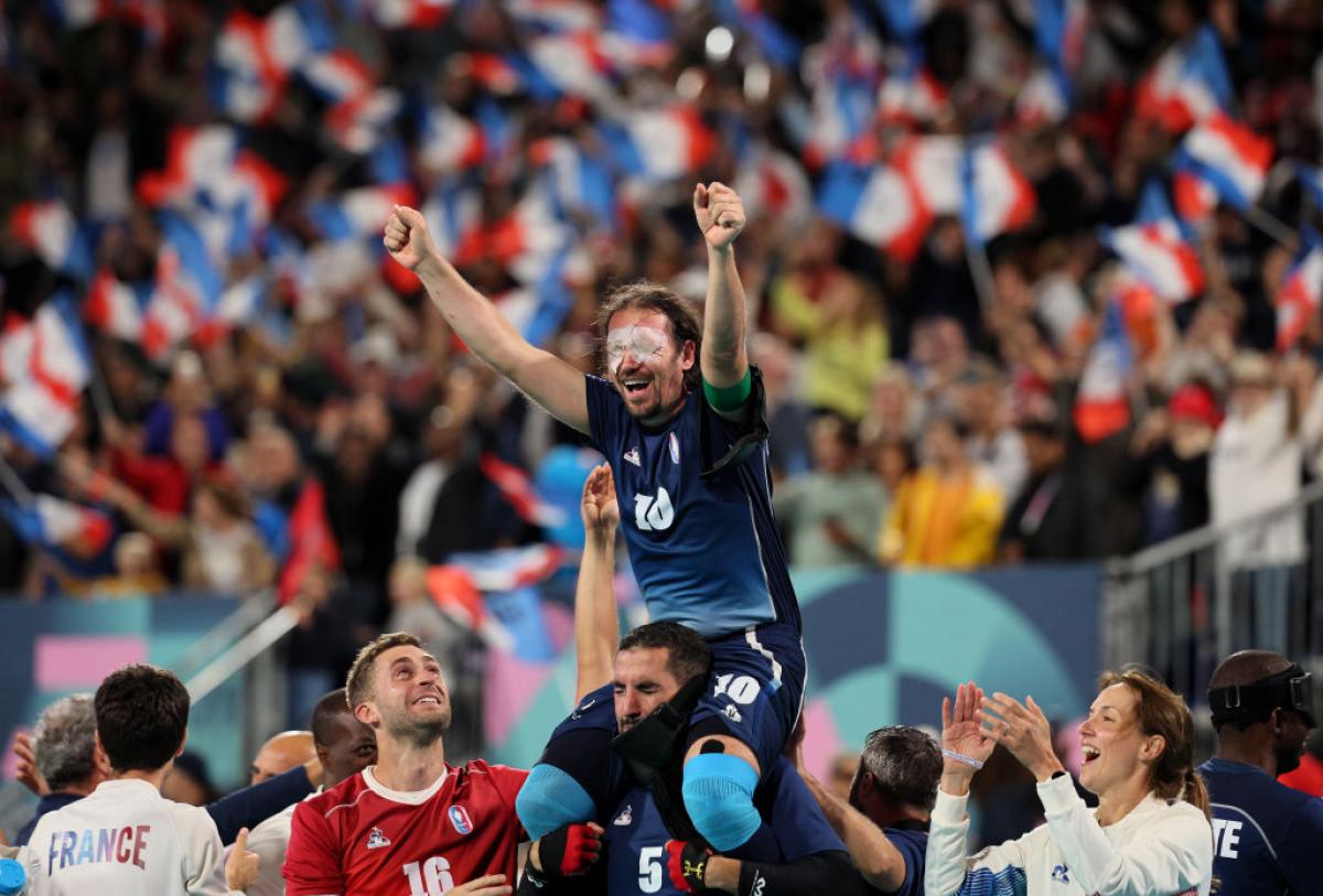 A man in a Team France football uniform with white patches over his eyes is lifted onto the shoulders of one of his teammates, as the crowd in the background waves French flags