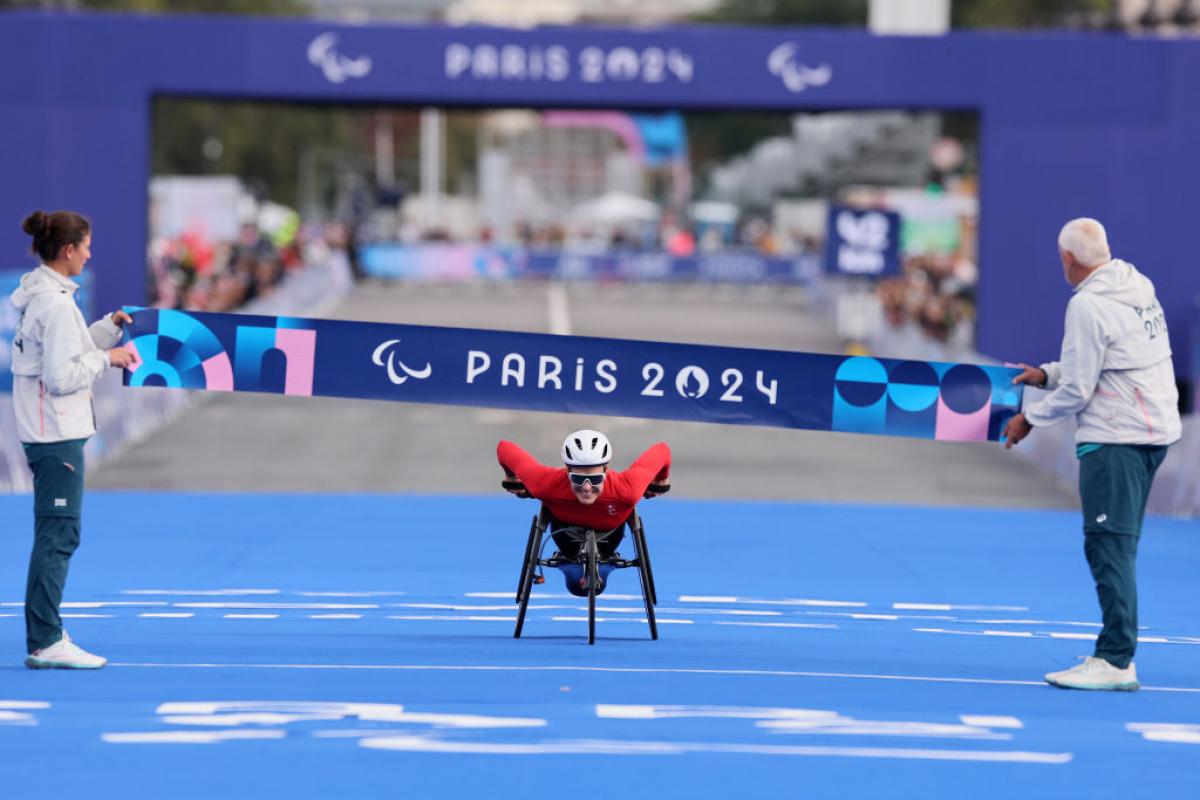 Woman in white helmet, sunglasses and red outfit leans forward in her racing wheelchair with a big smile as she approaches the finish line