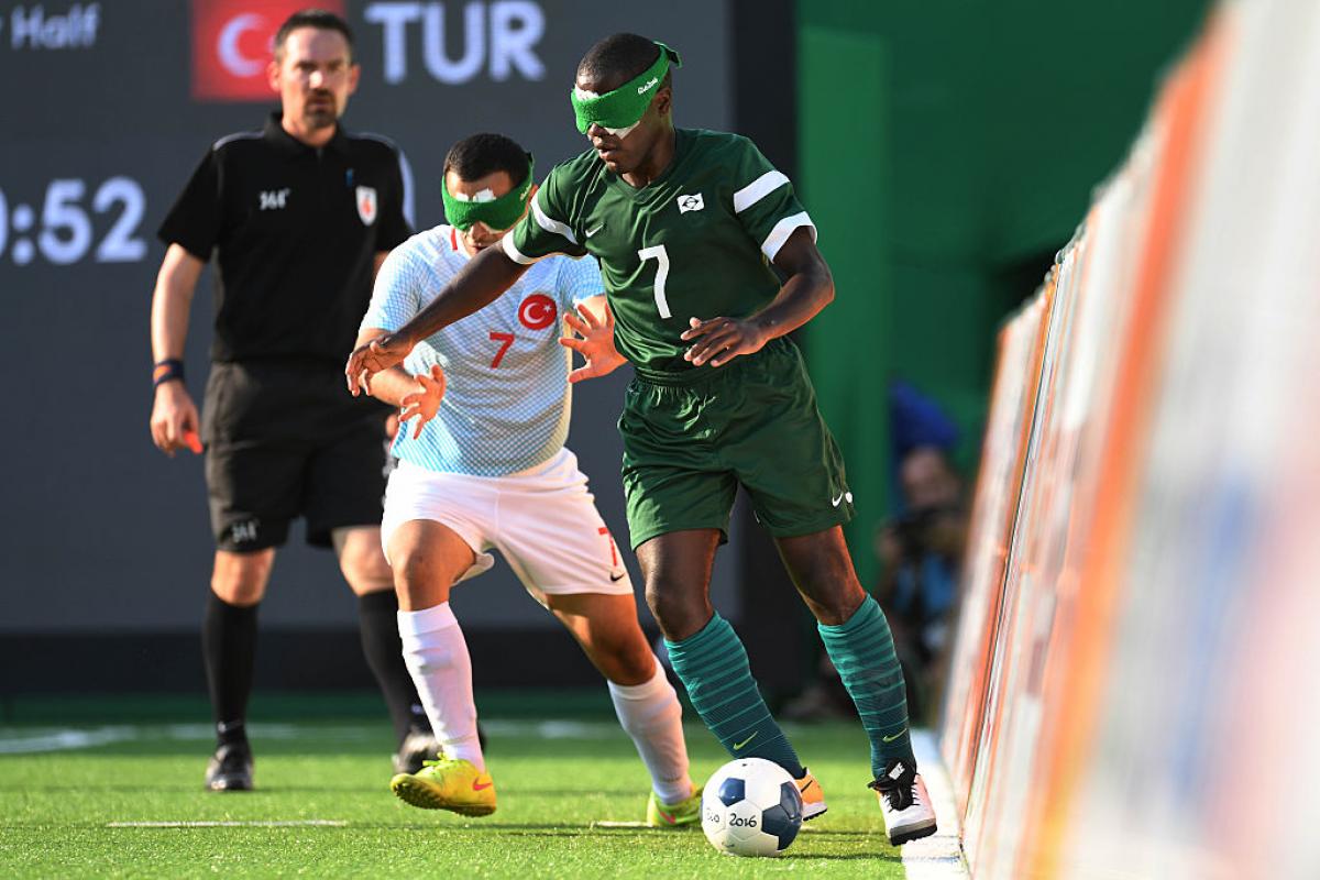 A man in green with a green blind fold tackles a man in blue taking the football away