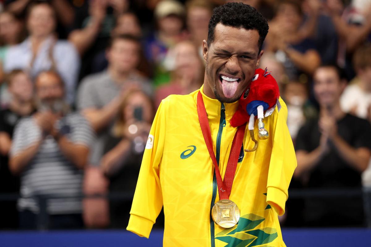 A male Para swimming athlete poses for a photo on the podium. He sticks his tongue