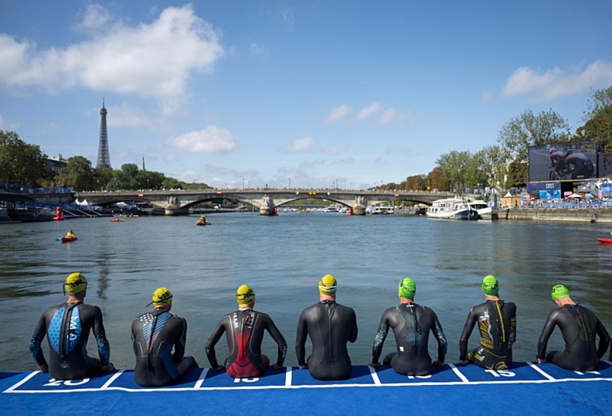 Six Para triathlon athletes are preparing for the swimming segment. The photo captures their back, sitting in front of a river