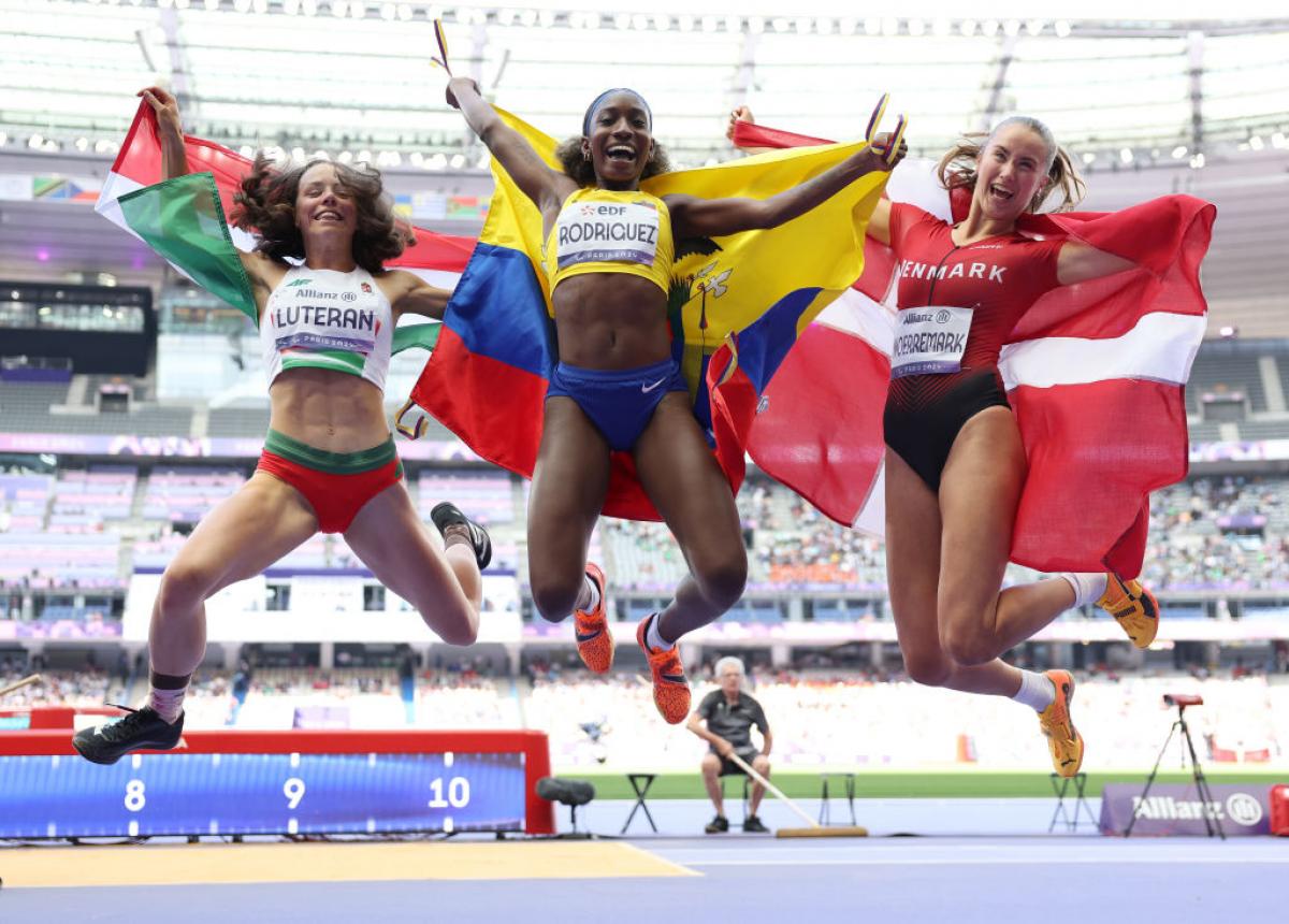 Three female athletes hold their flags and jump to celebrate 
