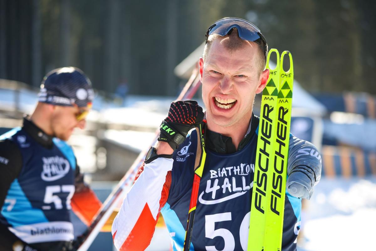 A male Para biathlon athlete smiles at the camera after competition. He is holding his skis