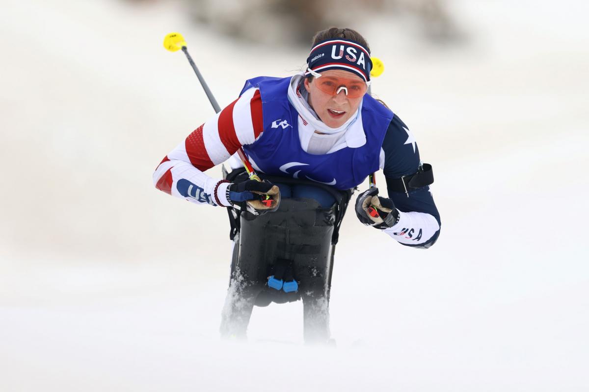 A female athlete competes in a sit-ski. She is wearing a head band that says USA and uniform in red, white and blue.